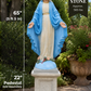 A cast stone statue of the Virgin Mary on a pedestal, shown outdoors in a garden setting. The statue features a blue robe and white gown, with a white head covering.