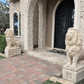 Beige concrete lion statue on square pedestal at entrance of stone house, with second lion visible in background