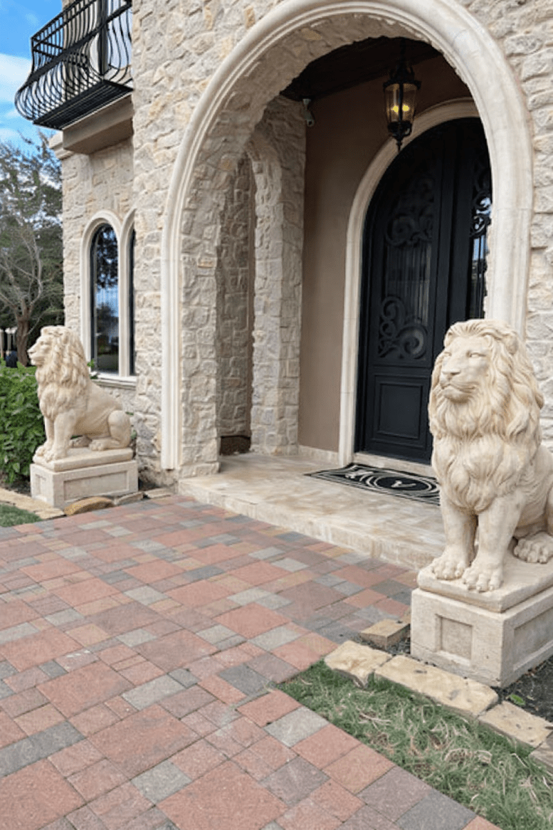Beige concrete lion statue on square pedestal at entrance of stone house, with second lion visible in background